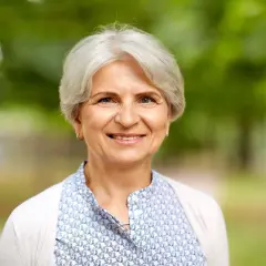 Uśmiechnięta starsza kobieta z siwymi włosami na tle zieleni.