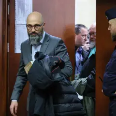 Mężczyzna w garniturze z książką i kurtką, obok policjant i inni mężczyźni w pomieszczeniu.