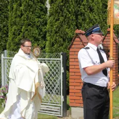 Ksiądz w białej szacie niesie monstrancję, obok stoją mężczyźni w mundurach.