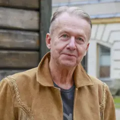 Bogusław Linda w brązowej kurtce z zamszu, z frędzlami i ozdobnymi szwami.