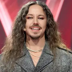 Michał Szpak w błyszczącej kurtce i z długimi włosami, uśmiecha się do kamery.