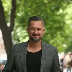 Marcin Prokop w szarej marynarce i czarnej koszulce, uśmiecha się na tle zieleni.