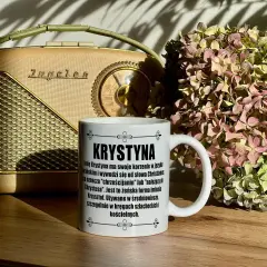 Biały kubek z napisem "Krystyna" na tle starego radia i bukietu suszonych hortensji.