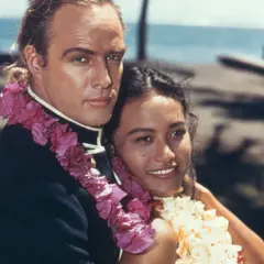 Marlon Brando z kobietą w kwiatowych girlandach na tle palmy i oceanu.