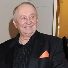Uśmiechnięty Bohdan Łazuka w czarnym garniturze z pomarańczową poszetką.