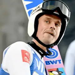 Piotr Żyła w kasku i goglach narciarskich, z nartami za plecami.