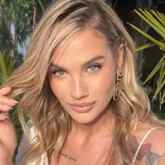 Karolina Gilon z blond włosami, niebieskimi oczami i tatuażem na ramieniu, na tle roślinności.