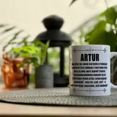 Biały kubek z napisem "ARTUR" i tekstem o znaczeniu imienia, na tle roślin.