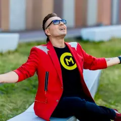 Czadoman w czerwonej marynarce i czarnej koszulce z logo siedzi na ławce z rękami rozłożonymi.
