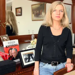 Kobieta z blond włosami w czarnej bluzce i jeansach, obok książka "Hołd Katyński" i ramka ze zdjęciem.