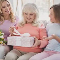Trzy kobiety na kanapie: starsza otwiera prezent, młodsza trzyma bukiet, najmłodsza gestykuluje.