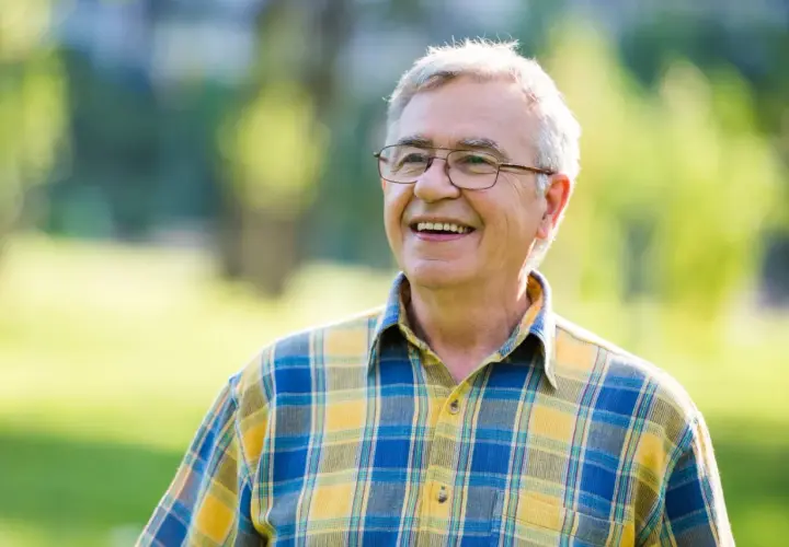 Uśmiechnięty starszy mężczyzna w okularach i koszuli w kratę na tle zieleni.