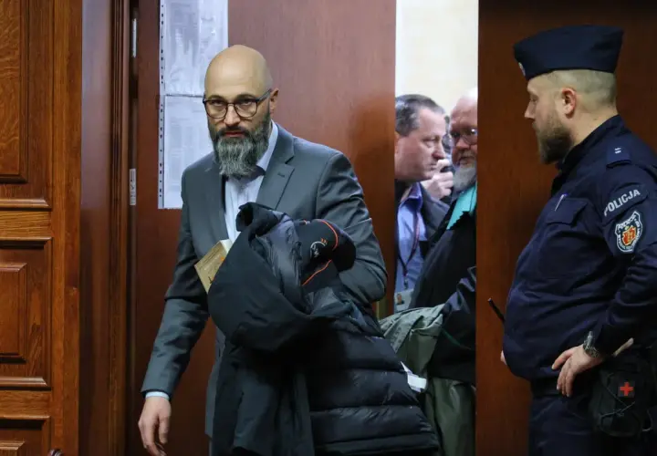 Mężczyzna w garniturze z książką i kurtką, obok policjant i inni mężczyźni w pomieszczeniu.