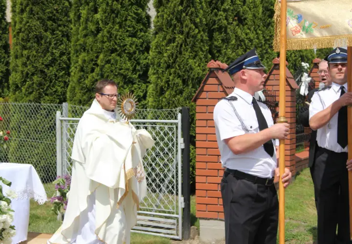 Ksiądz w białej szacie niesie monstrancję, obok stoją mężczyźni w mundurach.