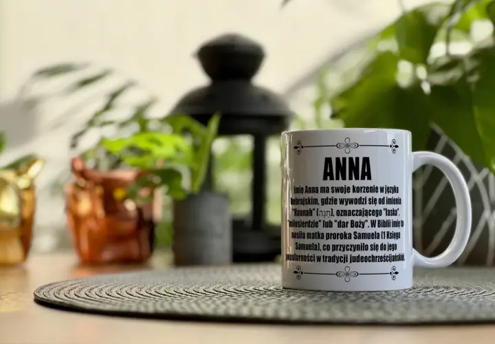 Biały kubek z napisem "ANNA" i tekstem o znaczeniu imienia, na tle roślin.