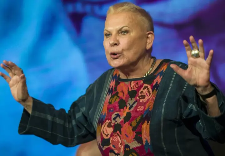 Elżbieta Dzikowska w kolorowej bluzce z haftem, gestykuluje rękami na tle niebieskiego ekranu.