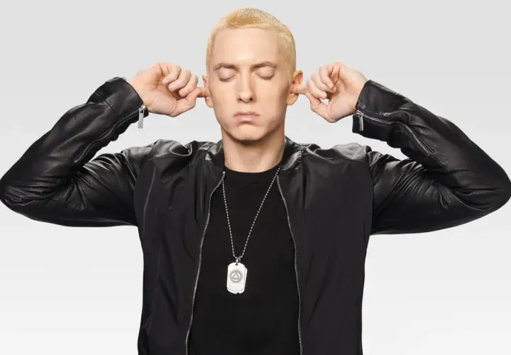 Eminem z zamkniętymi oczami zatyka uszy palcami, ubrany w czarną kurtkę.