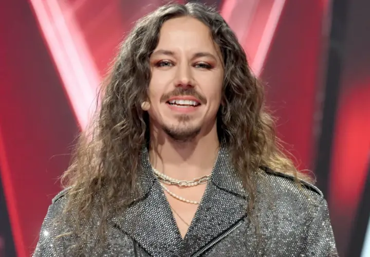 Michał Szpak w błyszczącej kurtce i z długimi włosami, uśmiecha się do kamery.