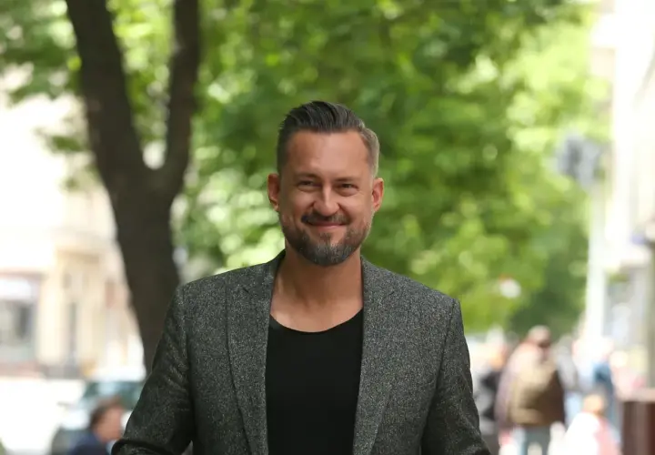 Marcin Prokop w szarej marynarce i czarnej koszulce, uśmiecha się na tle zieleni.