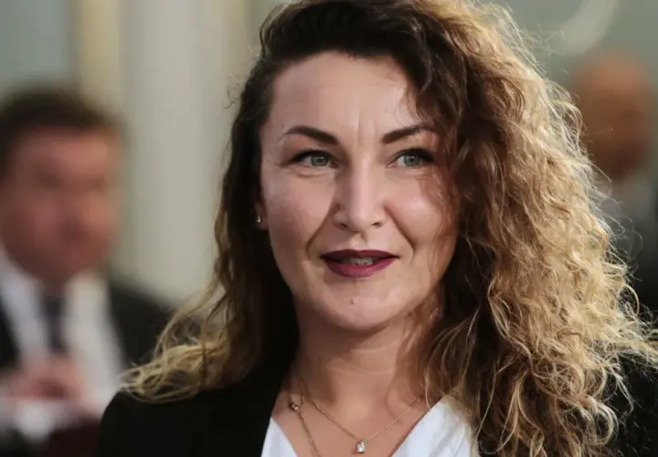 Monika Pawłowska z kręconymi włosami, w białej bluzce i czarnej marynarce, uśmiecha się.