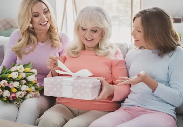 Trzy kobiety na kanapie: starsza otwiera prezent, młodsza trzyma bukiet, najmłodsza gestykuluje.