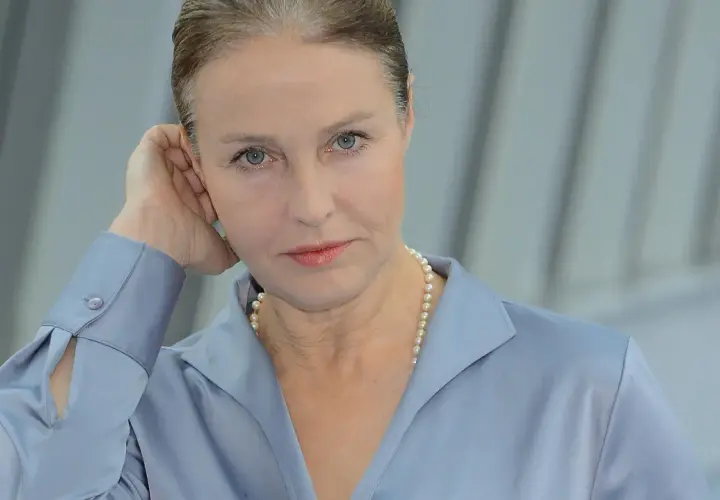 Portret kobiety w niebieskiej koszuli i perłach, opierającej dłoń o ucho.