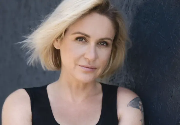 Małgorzata Ohme z blond włosami i tatuażem na ramieniu, na ciemnym tle.
