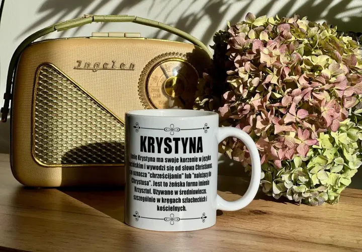 Biały kubek z napisem "Krystyna" na tle starego radia i bukietu suszonych hortensji.