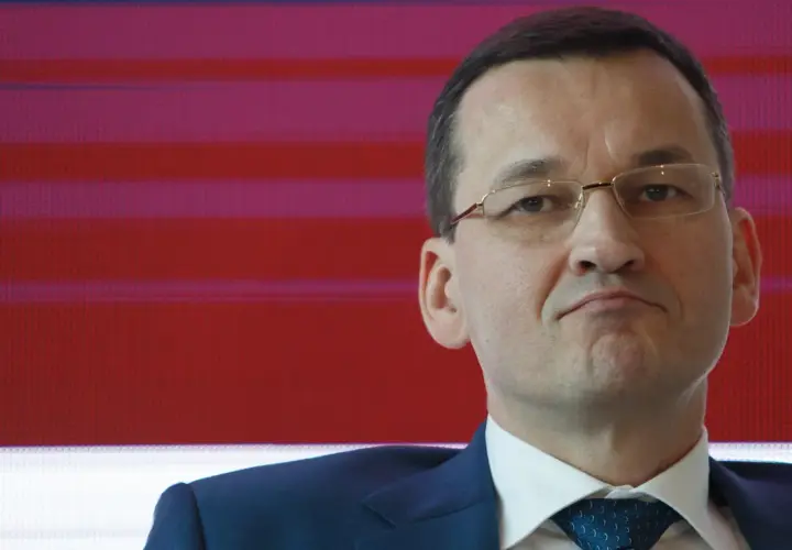 Mateusz Morawiecki w garniturze i okularach na tle czerwono-fioletowych pasów.