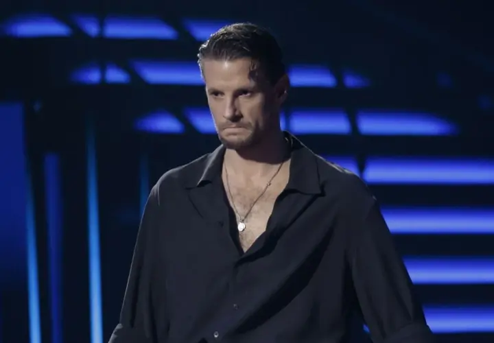 Sebastian Fabijański w czarnej koszuli z naszyjnikiem na tle niebieskich świateł.