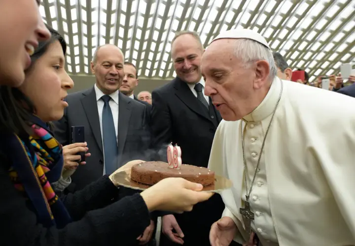 Papież Franciszek zdmuchuje świeczki na torcie urodzinowym, trzymanym przez kobietę.