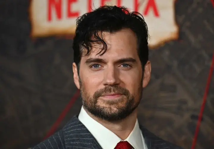 Henry Cavill w garniturze i czerwonym krawacie, z zarostem i niebieskimi oczami.
