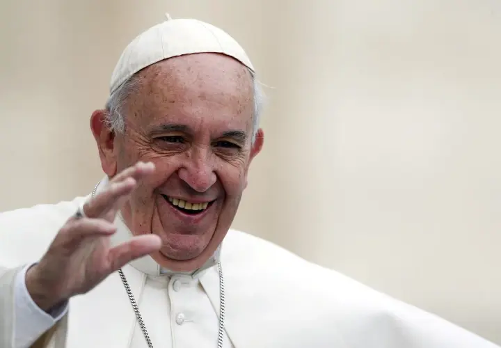 Papież Franciszek w białej sutannie i piusce, uśmiechnięty, macha ręką.