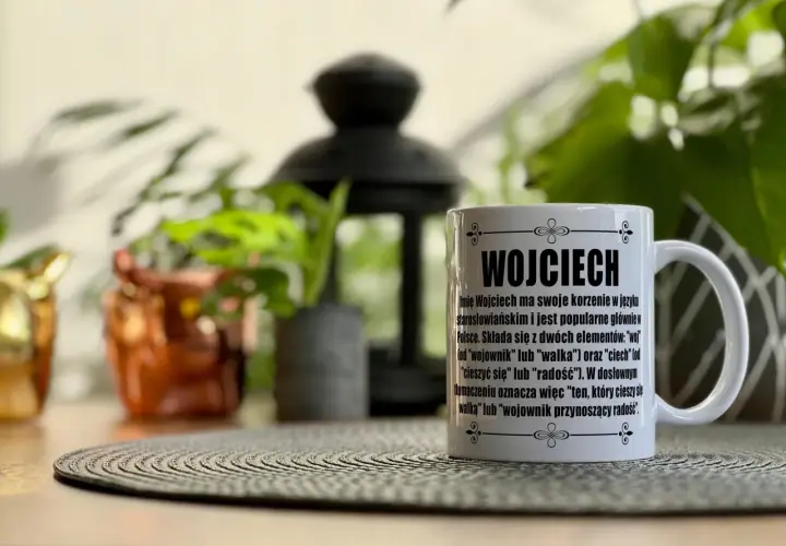 Biały kubek z napisem "Wojciech" i opisem znaczenia imienia, na tle roślin.