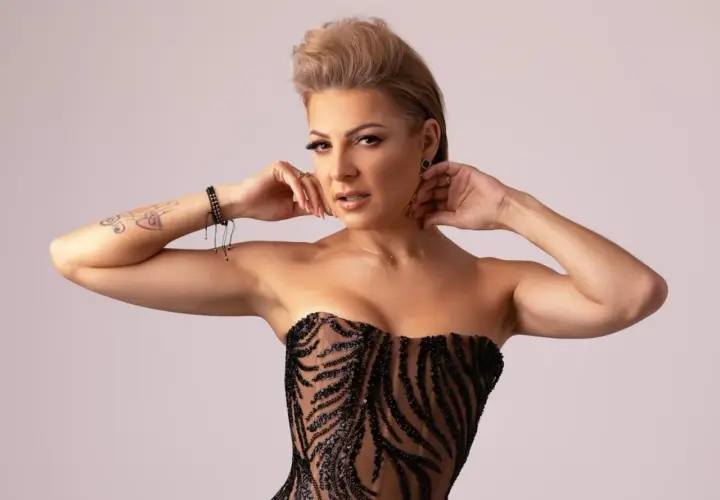 Magda Narożna w czarnej, błyszczącej kreacji, z dłońmi przy twarzy i tatuażem na ramieniu.