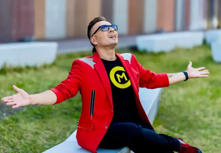Czadoman w czerwonej marynarce i czarnej koszulce z logo siedzi na ławce z rękami rozłożonymi.