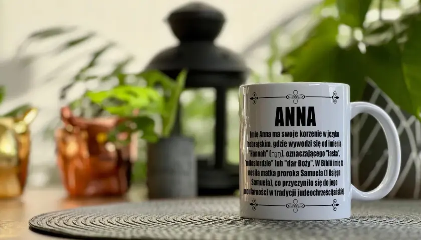 Biały kubek z napisem "ANNA" i tekstem o znaczeniu imienia, na tle roślin.