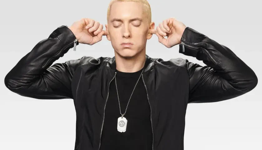 Eminem z zamkniętymi oczami zatyka uszy palcami, ubrany w czarną kurtkę.
