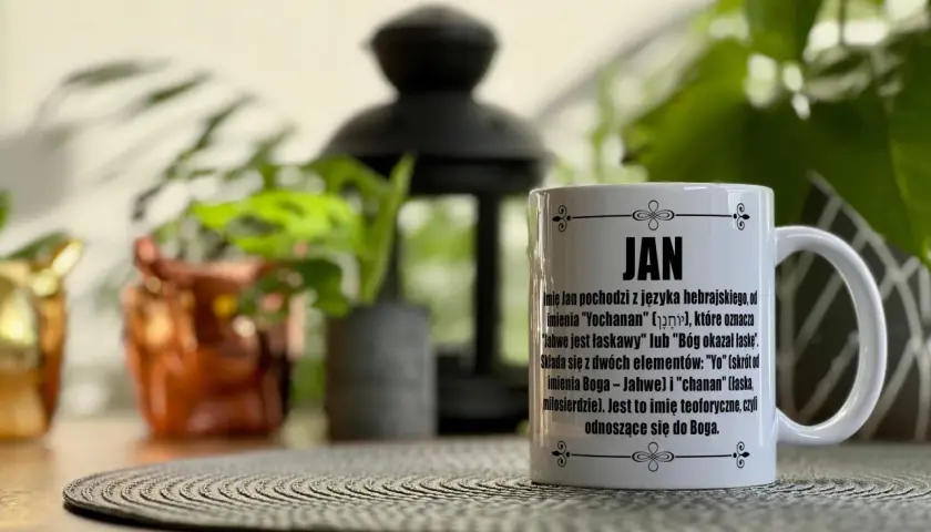 Biały kubek z napisem "JAN" i tekstem o pochodzeniu imienia, na tle roślin.