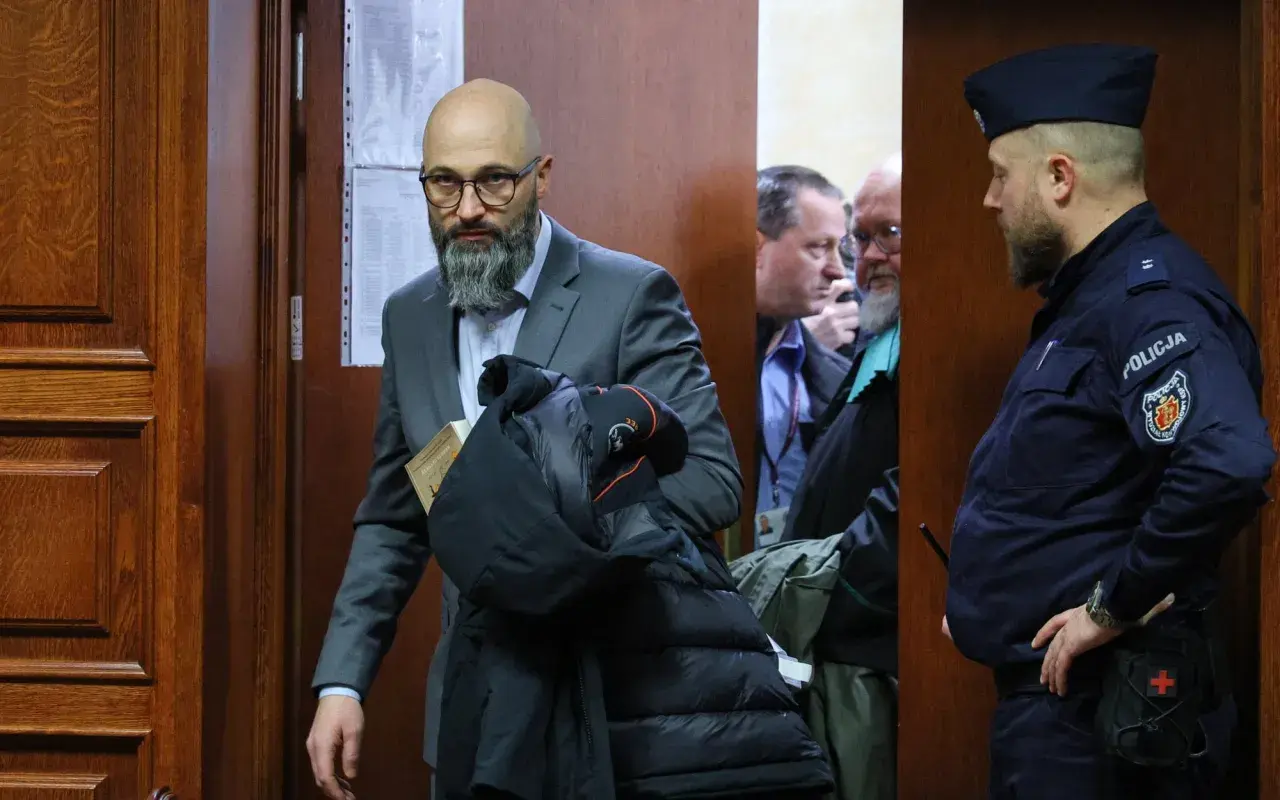 Mężczyzna w garniturze z książką i kurtką, obok policjant i inni mężczyźni w pomieszczeniu.
