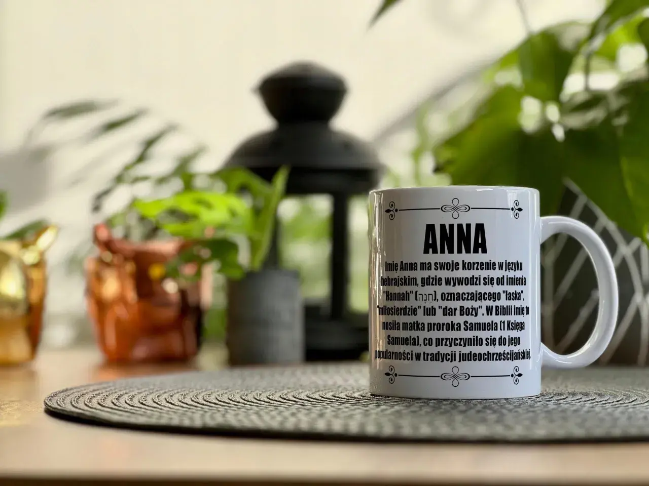 Biały kubek z napisem "ANNA" i tekstem o znaczeniu imienia, na tle roślin.
