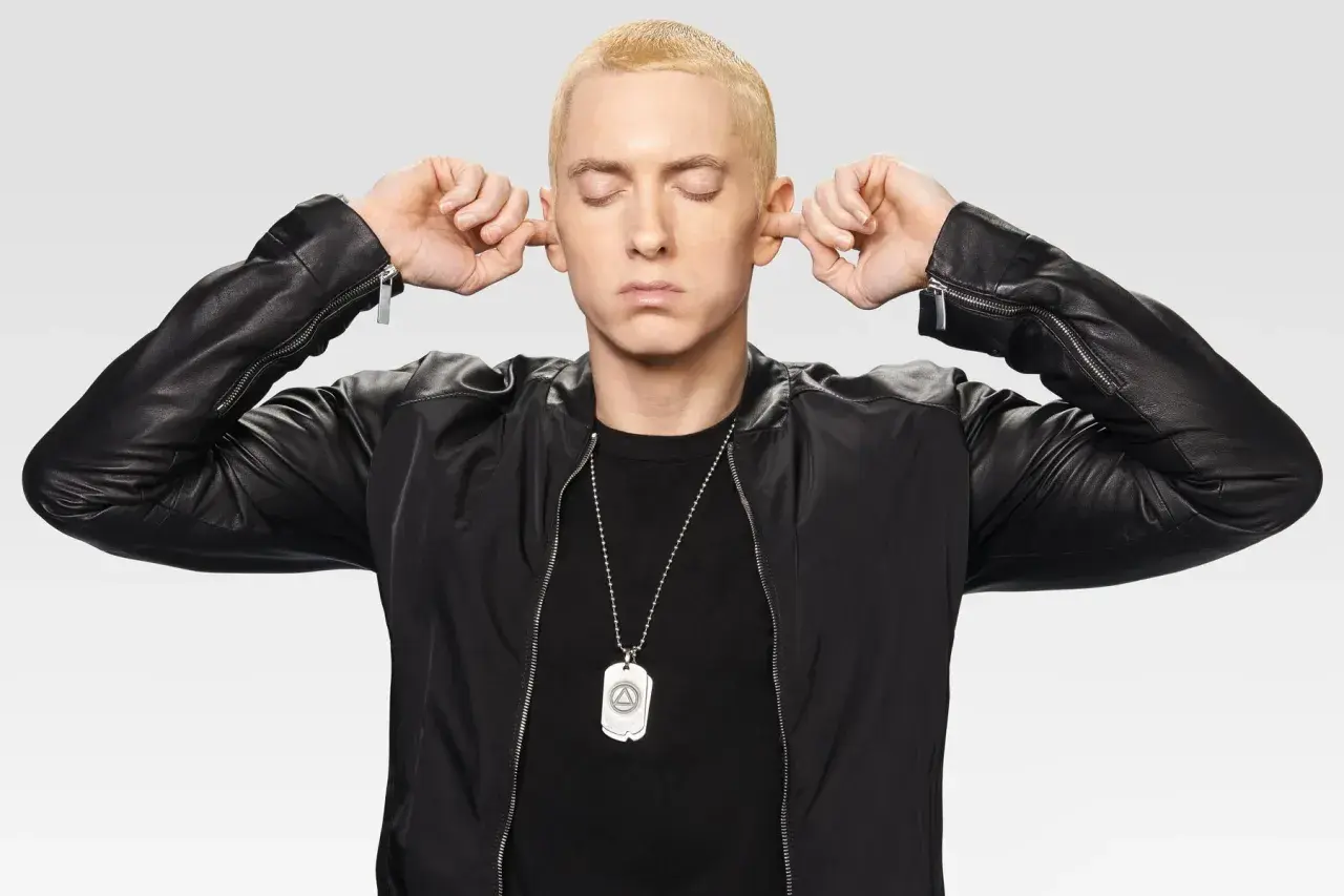 Eminem z zamkniętymi oczami zatyka uszy palcami, ubrany w czarną kurtkę.