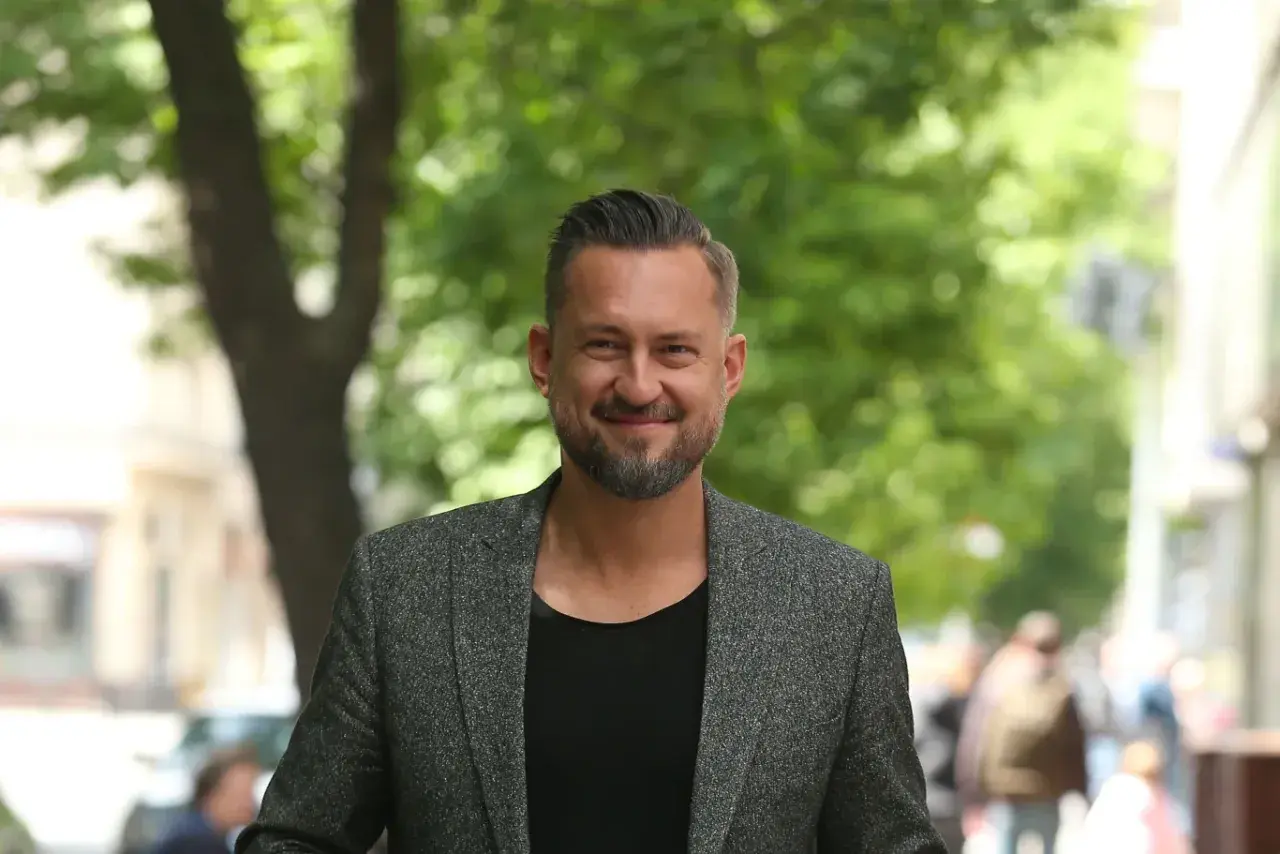 Marcin Prokop w szarej marynarce i czarnej koszulce, uśmiecha się na tle zieleni.