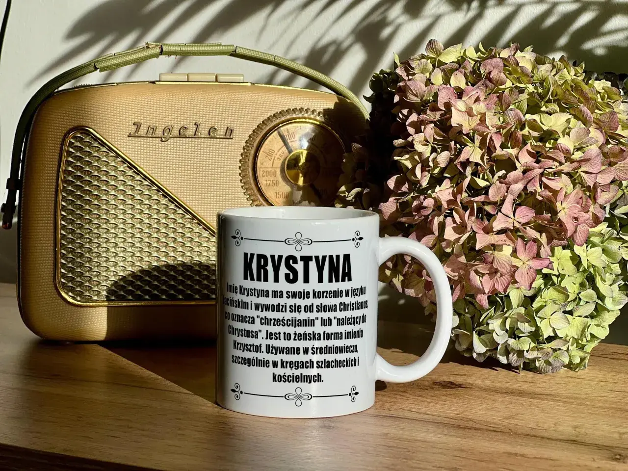 Biały kubek z napisem "Krystyna" na tle starego radia i bukietu suszonych hortensji.