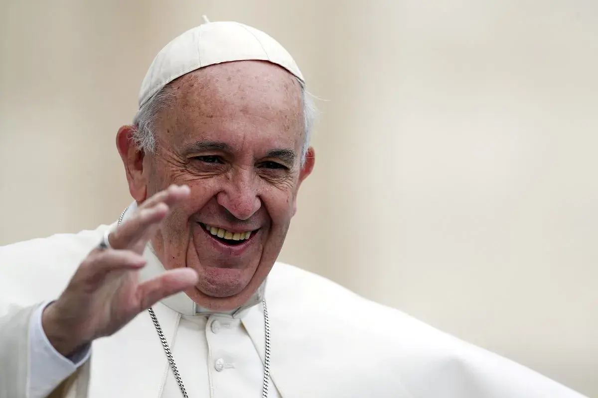 Papież Franciszek w białej sutannie i piusce, uśmiechnięty, macha ręką.