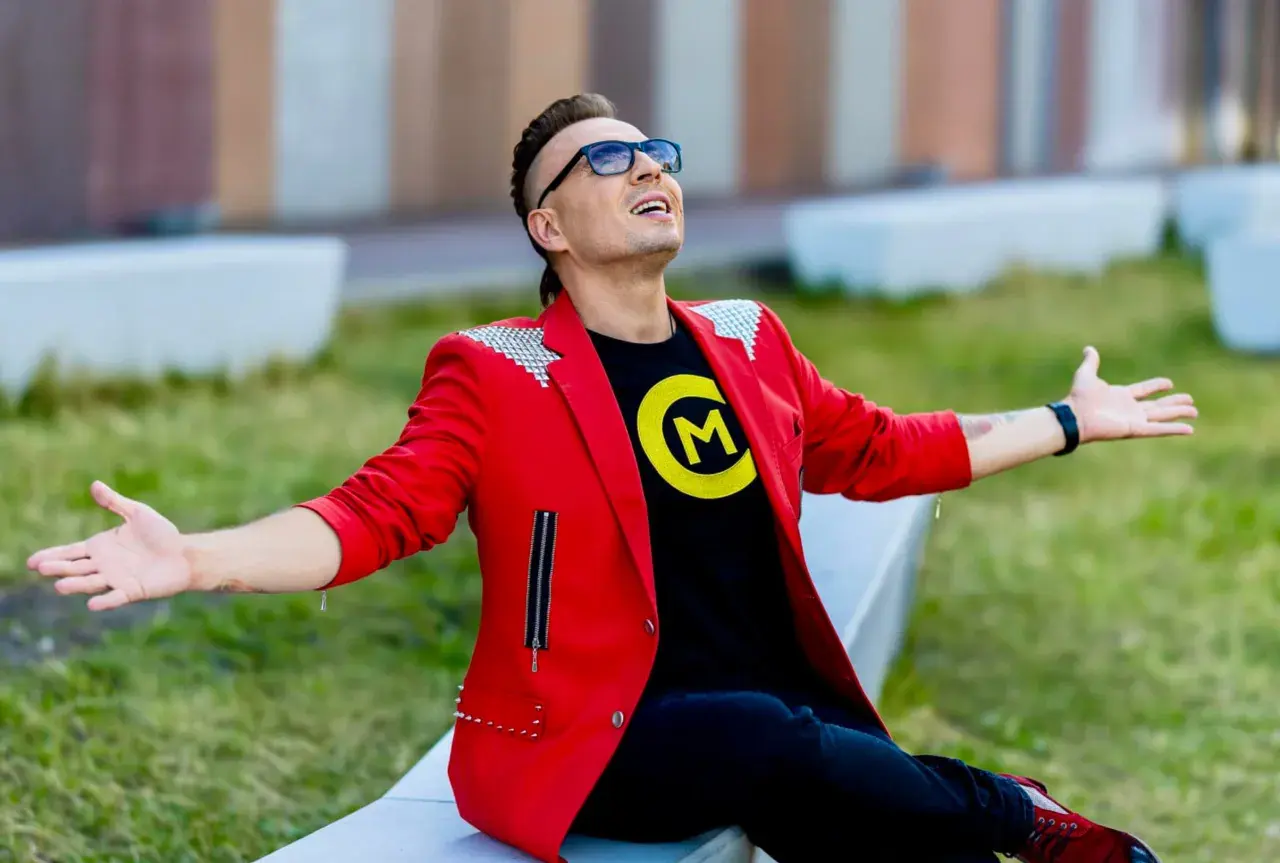 Czadoman w czerwonej marynarce i czarnej koszulce z logo siedzi na ławce z rękami rozłożonymi.